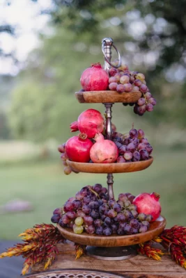 Three-tier rustic wooden fruit display with pomegranates and grapes, styled for a wedding or event catering table.