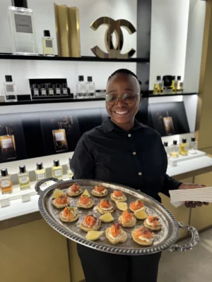 Smiling server presenting a silver tray of gourmet salmon canapés with lemon wedges at a luxury corporate event inside a Chanel boutique.