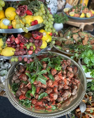 Large silver platter of Italian-style meatballs in tomato sauce with basil, served alongside a colorful tiered fruit display of lemons, grapes, pomegranates, and strawberries at a catered event.